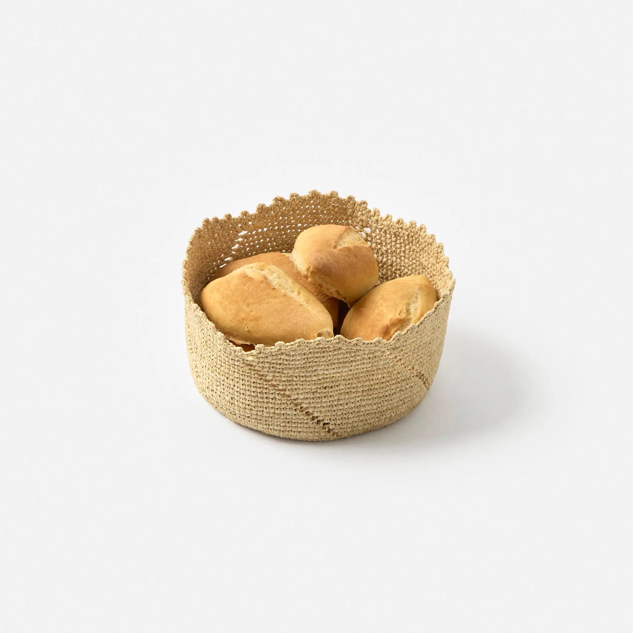 A round basket, crocheted with natural raffia, with small bread rolls inside, on a light grey background.